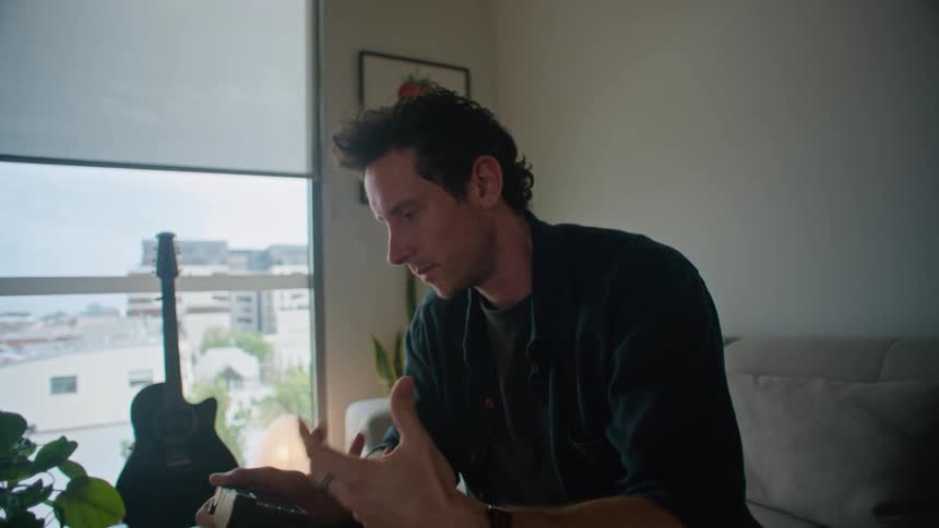 Man in black shirt and pants sits on gray couch and makes a video call on laptop while holding camera in the living room