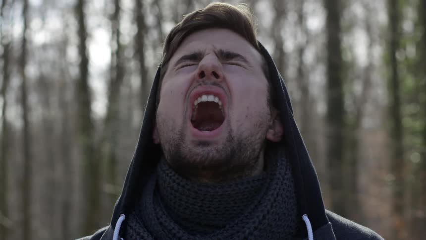Man wearing a hooded jacket tilts his head back and yells as he stands in a forest