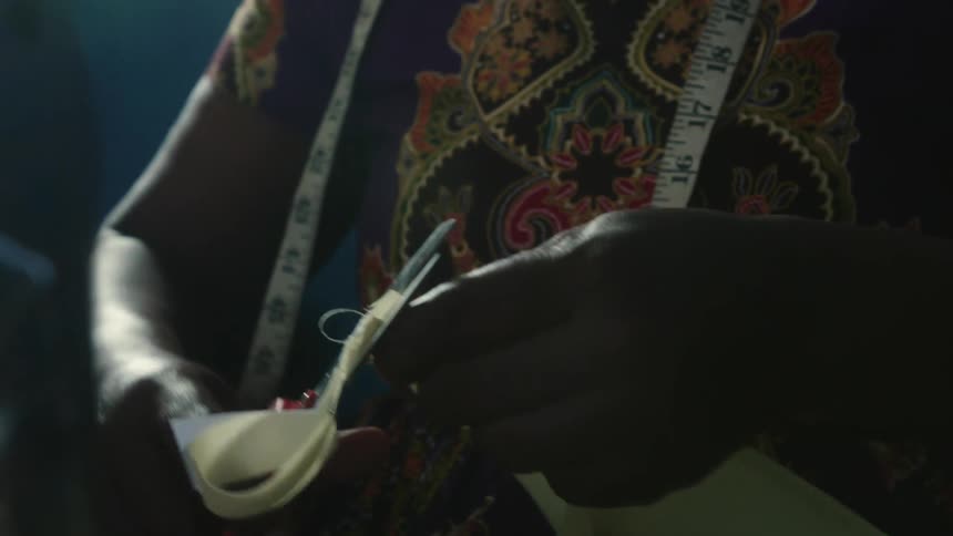 A woman standing near a sewing machine with a tape measure around her neck is holding a pair of scissors as she cuts a piece of fabric