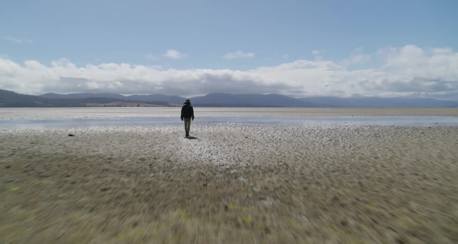 Man walks across sandy ground filled with multiple shallow puddles with misty mountains on the horizon