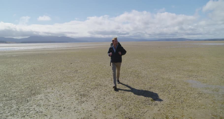 Man with backpack black jacket and brown pants walks on dry lake bed