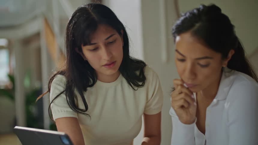 Two women are standing over a table with a computer as they look over plans and point as they talk