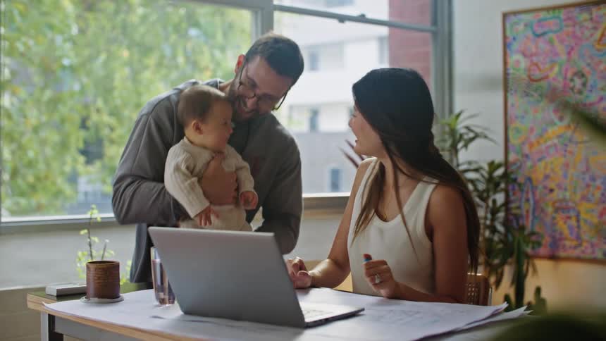 A woman is sitting at a desk working as her husband brings their baby for a visit
