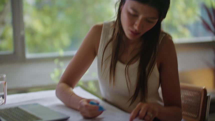 A woman is sitting at a desk with a laptop computer as she writes on blueprints with a marker
