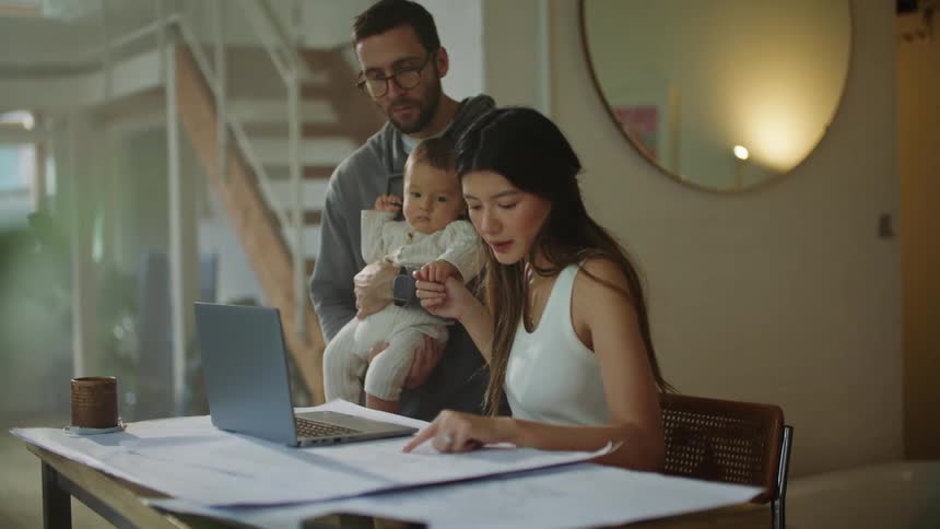 Mom and dad at home with baby mom is working on blueprint on drafting table with laptop
