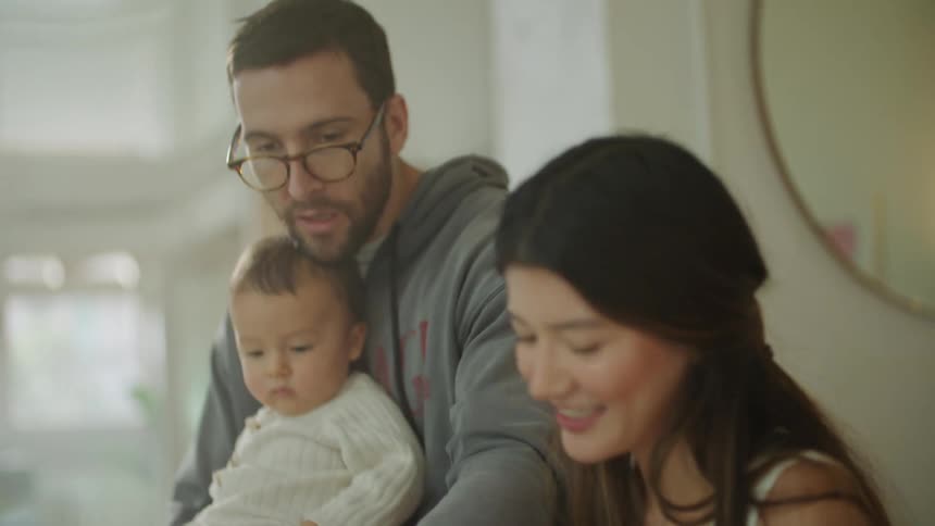 A woman sitting at a table with papers and a laptop in front of her while laughing at her husband as he holds their baby and they talk about the plans on paper
