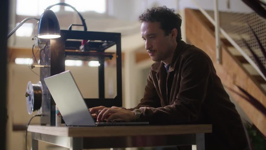 A man monitors a three dimensional printer while using his laptop