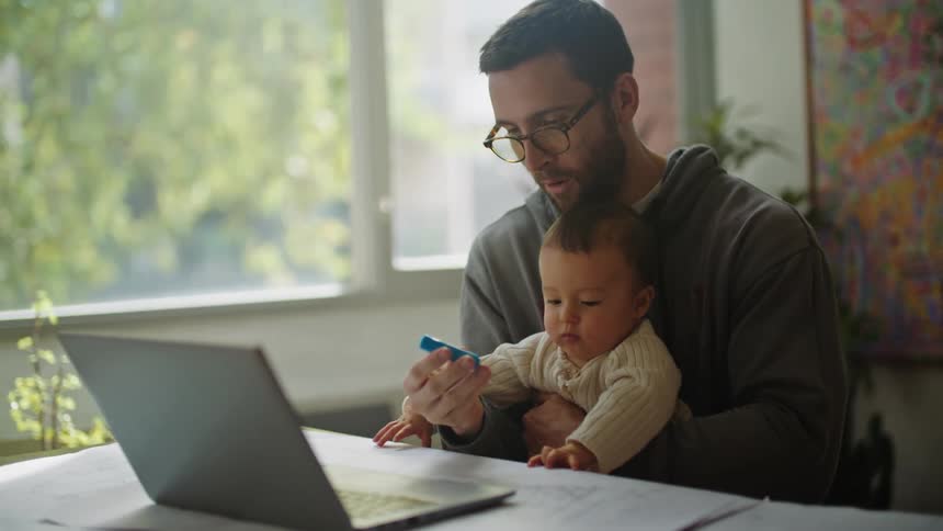 Father holds and smiles at child while working in home office