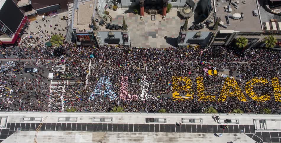 A crowd and parade of people on a street spell out the words all black using color