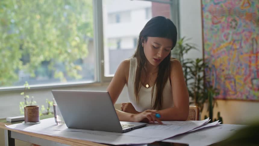 Woman with brown hair white shirt and necklace works on plans and laptop at the office