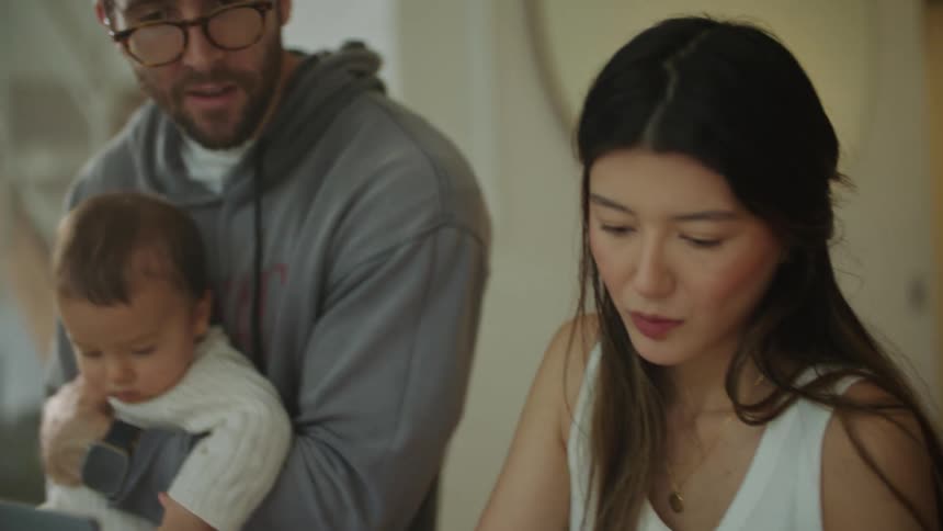 A man holding a baby in his arms talking to his wife sitting at a desk looking over papers under her laptop