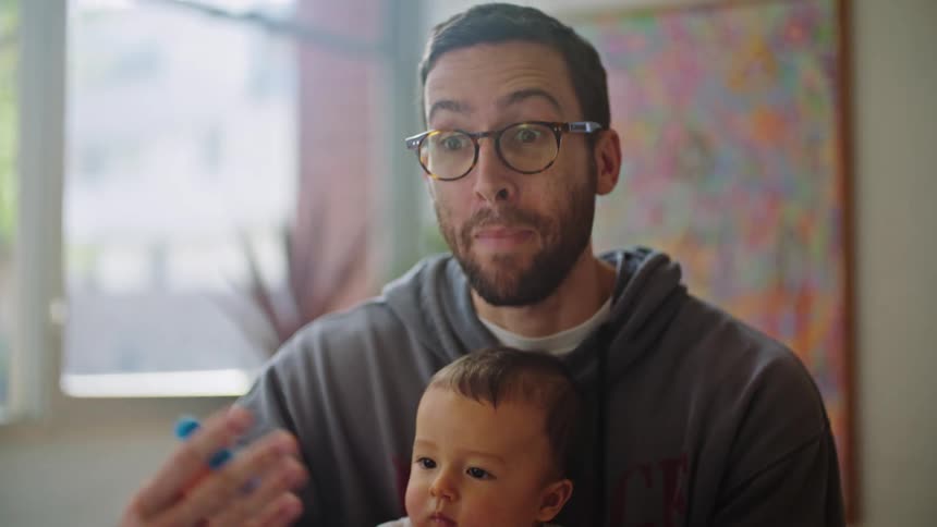 A man sitting in front of a laptop talking to it while holding his infant son on his lap