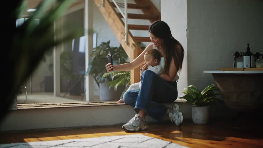 A woman holding her baby sitting on a large bay window sill posing for a selfie the mother is taking with her phone