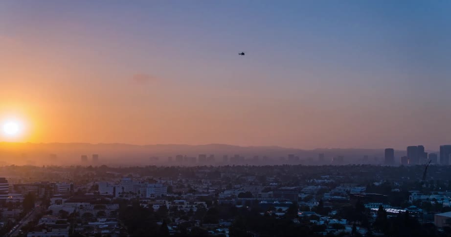 An aerial of a city skyline with the sun sitting on the horizon and a helicopter flying over the city