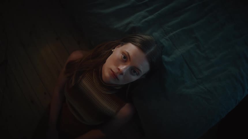 Woman with brown hair earrings and brown top looks around and at ceiling leaning against the bed in bedroom