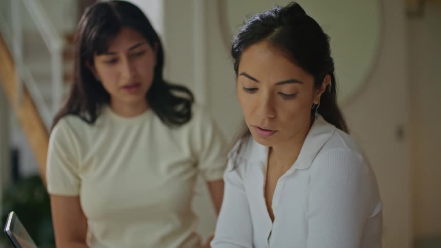 Two women talking and thinking while looking at plans on paper and then looking at the laptop in front of them
