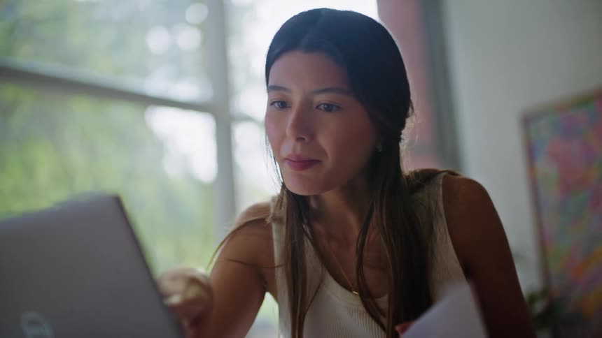 Young woman sitting at desk in home office looking at laptop screen and referencing paperwork