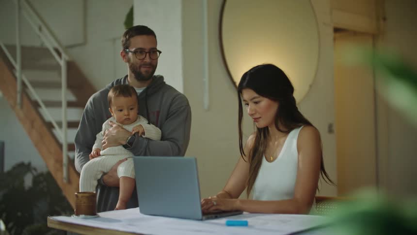 A woman sitting at a desk in front of her laptop while her husband and baby stand next to her as she interacts with them