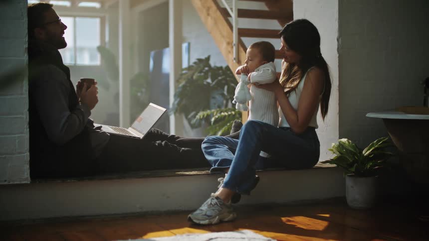 A mother holds her baby up right facing the dad and husband sitting across from each other with a laptop on his lap