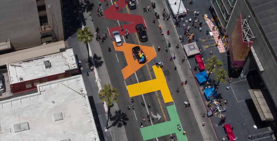 A crowd follows behind a vehicle driving over an art installation on a city street