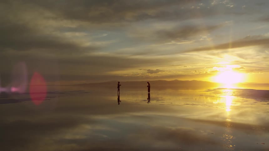 Two silhouettes stand on a beach at sunset