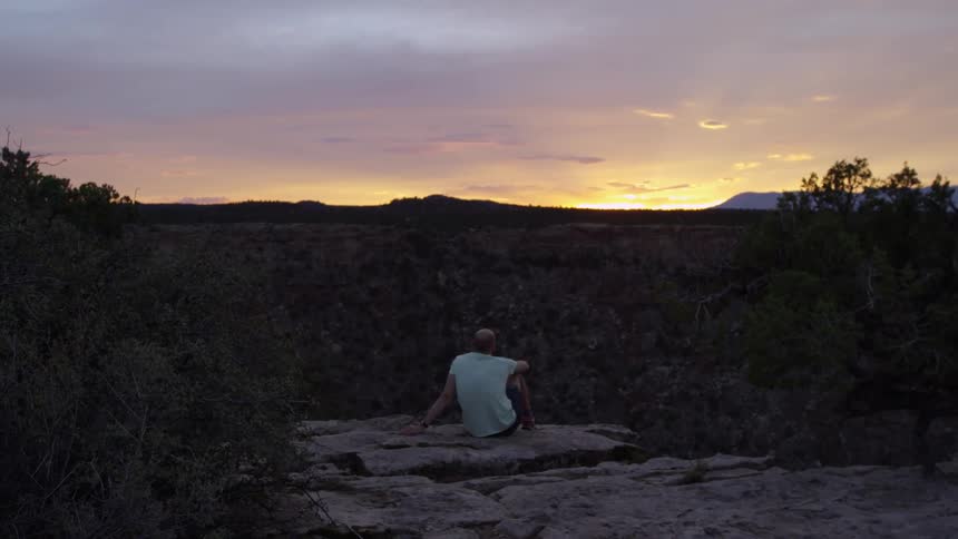 Man sits on cliff overlooking trees at sunset