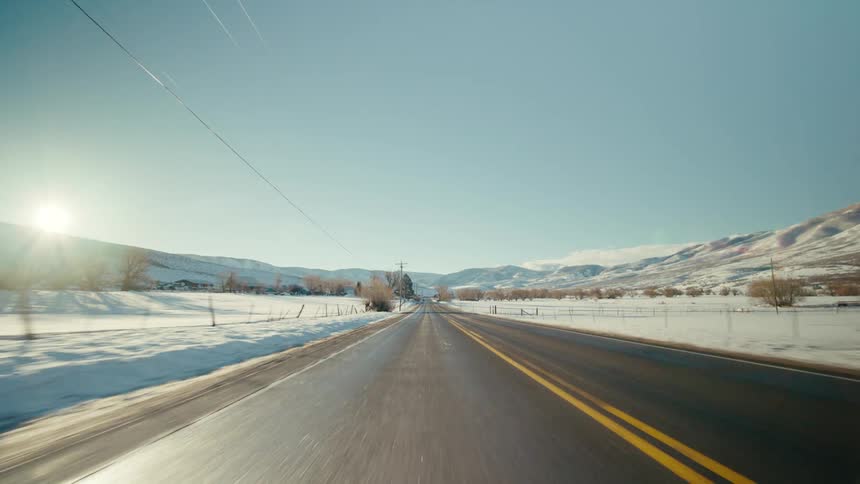 Traveling on an open rural road with mountains in the distance and snow on the roadside as the sun shines brightly on a winter day