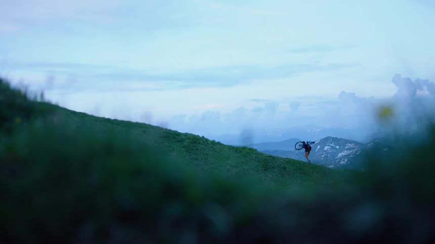 Distant biker goes up green mountain slope carrying bicycle at dusk