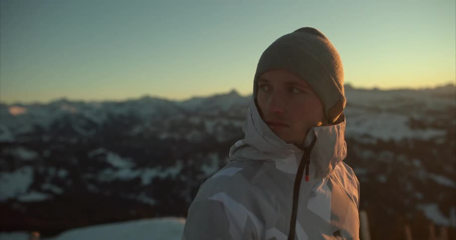 Hiker with wooly hat and gray coat takes photos with camera on snowy mountain at sunset