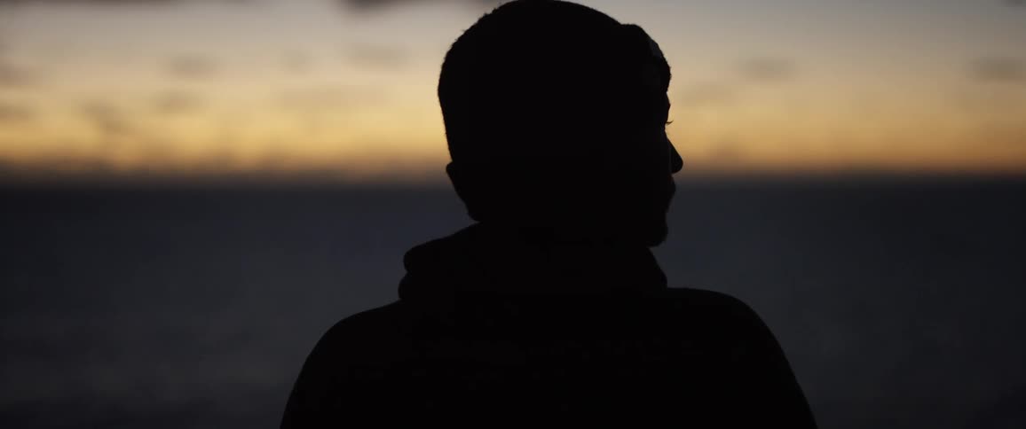 Man with wooly hat and jacket is silhouetted against the sky at sunset watching the ocean