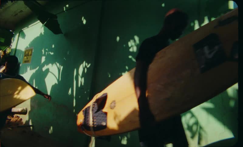 A group of boys walk single file with their surf boards next to a green building with shadows on it