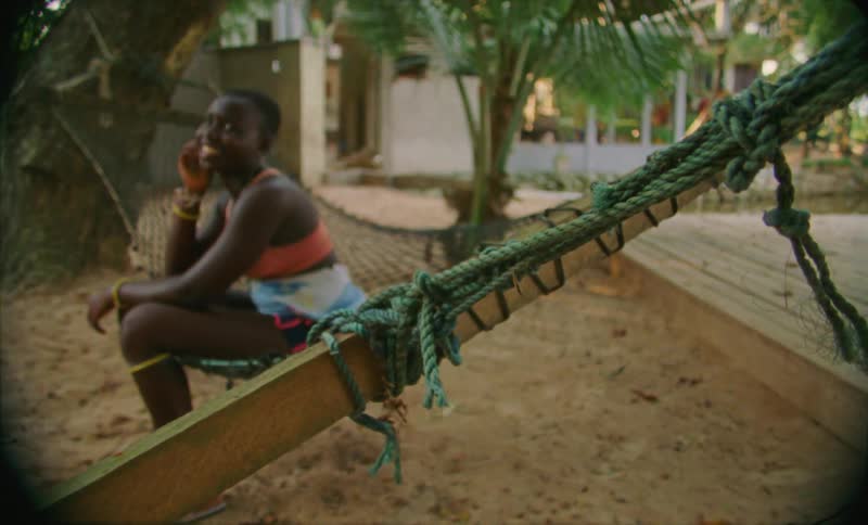 An african woman sits on a hammock and talks to someone unseen