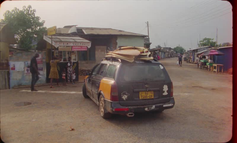 Car with several passengers and surfboards on roof turns onto street passing pedestrians