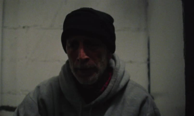 A man wearing a knit cap and a gray hoodie drinks from a whiskey bottle