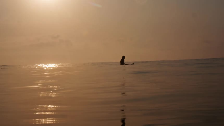 A lone surfer floats on a surfboard in the middle of the ocean at dusk