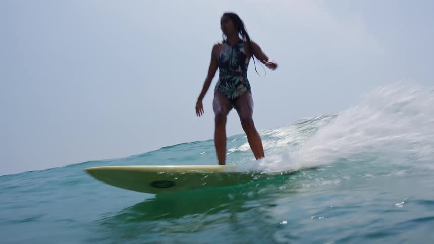A teenage girl is standing on her surfboard moving her arms and keeping her balance as she rides a wave in to shore