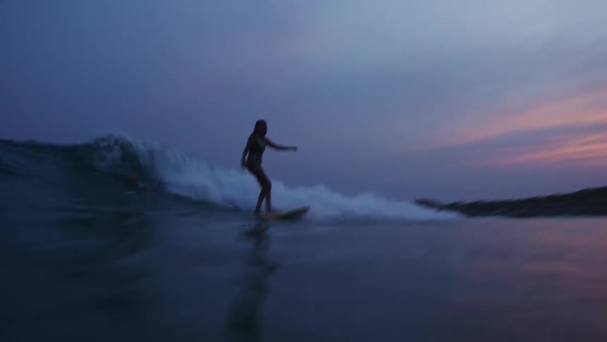 A time lapse of a silhouette of a woman riding a surfboard on a wave in the ocean at dusk