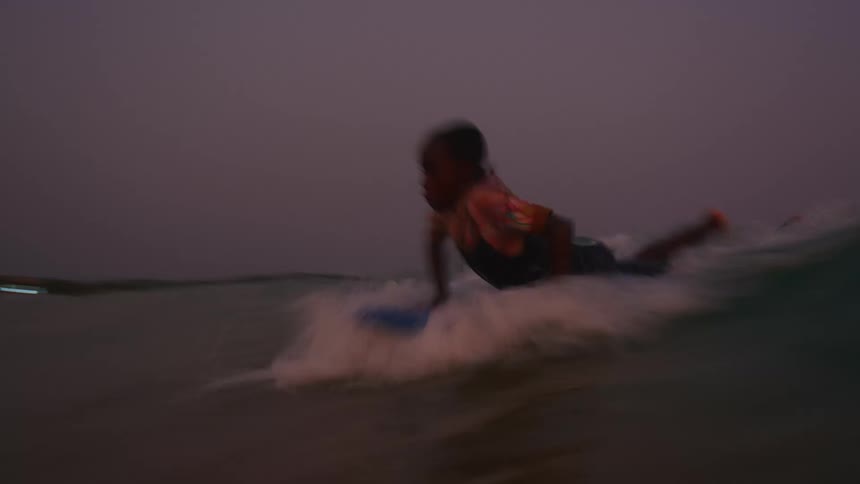 A young boy climbs atop his surfboard and rides a wave to the shore at sunset