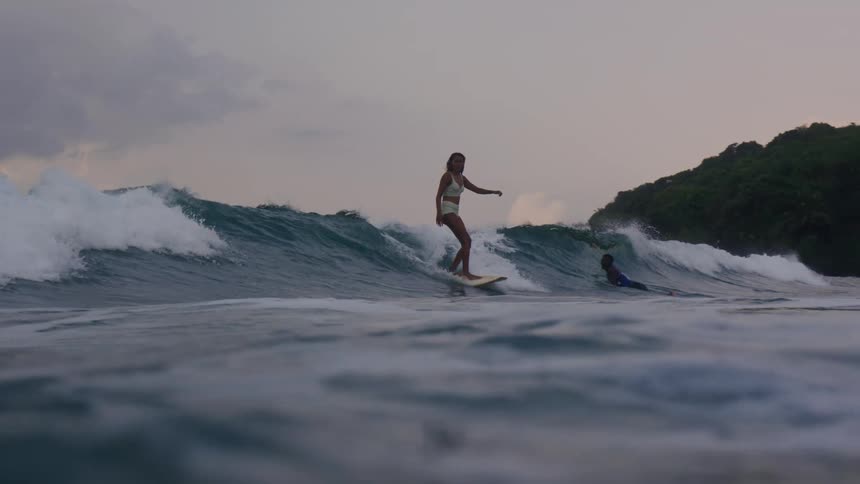 A woman on a surfboard rides a wave as a man swims nearby in the sea at dusk