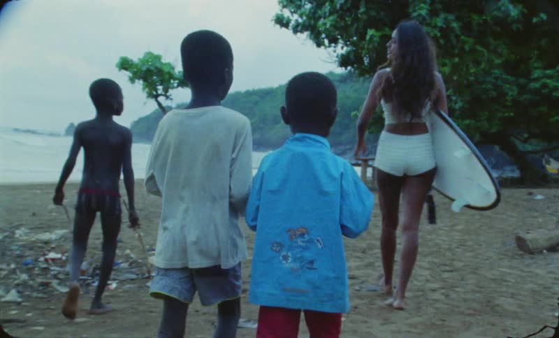A woman with a surfboard and three children walking along the beach on an island