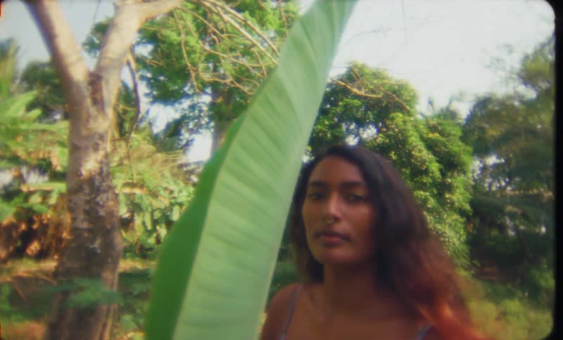 Young woman stands near a the large leaf of a tropical plant at morning caught on vintage film