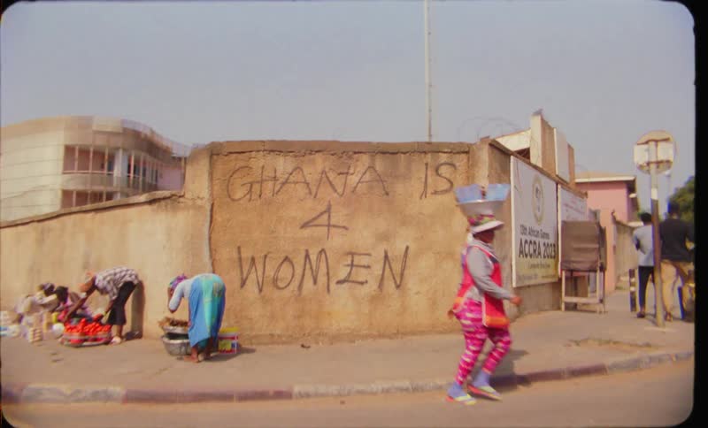 Vehicle crosses intersection as street merchant bends down near wall with the slogan ghana is 4 women