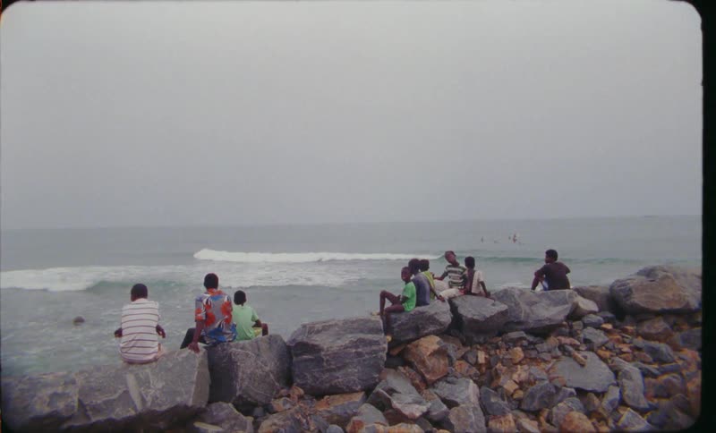 Youths sit on boulders at beach looking out to sea as waves roll toward shore