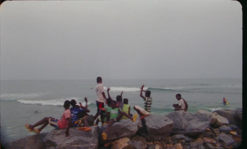 A group of young black boys sit on stones near the ocean shore and play