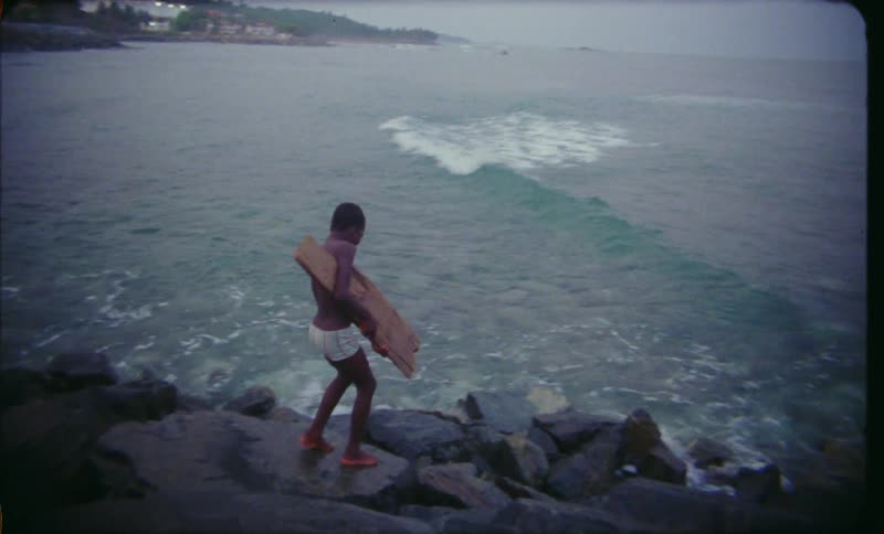 A boy is carrying a board as he walks on rocks to the water along the coast