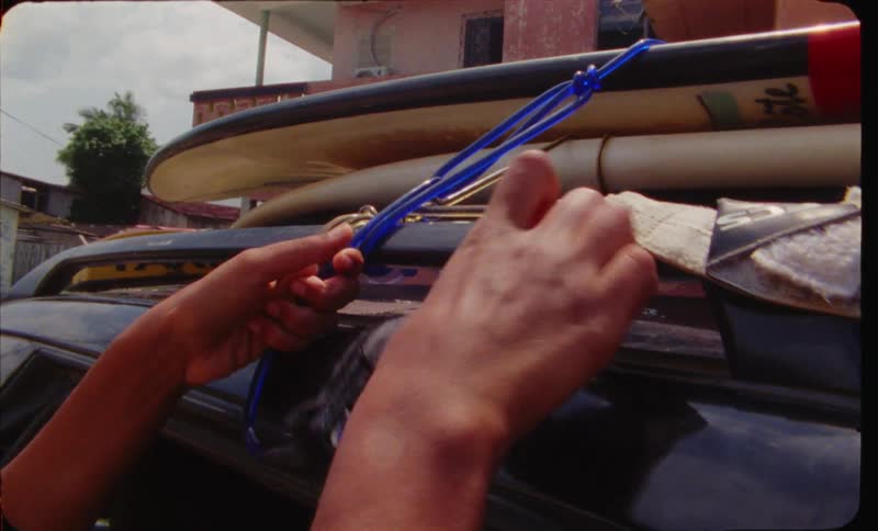 A woman straps a surfboard to the top of her vehicle as a man looks on