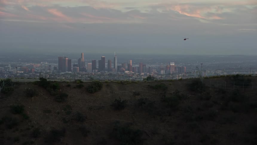An epic aerial footage shot during twilight in the summer showing Los Angeles downtown skyline from above as the camera ascends over a ridge