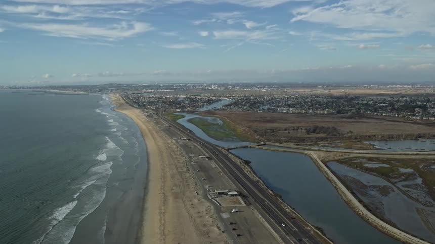 Aerial footage of Bolsa Chica State Marine Conservation Area in Huntington Beach CA showcasing the coastline and Highway 1 with light traffic on a partly cloudy day during summer
