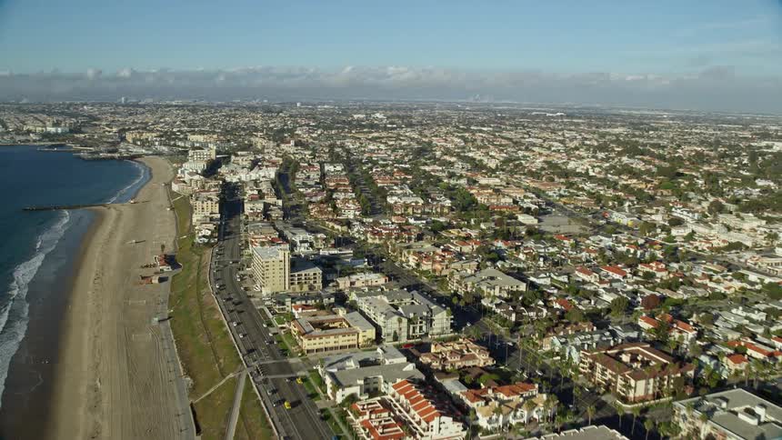 Aerial footage of Torrance Beach in Redondo Beach CA approached from the west coast with high wide-angle shot over an empty beach and shoreline with partly cloudy skies amidst residential and coastal communities