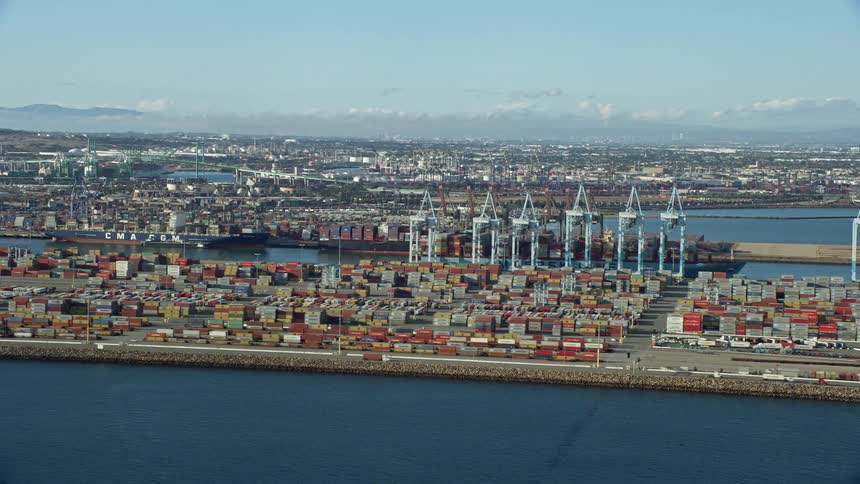 An epic aerial view of the Port of Los Angeles with shipping containers and cargo cranes set against a partly cloudy sky and the cityscape in the background